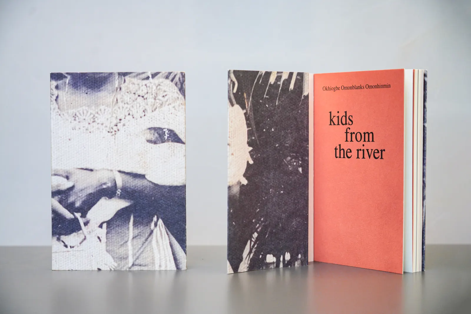 Two copies of kids from the river on a grey table. One is closed, showing the cover photograph of the author's mother holding her first child in 1979. The other stands open at the coral title page.